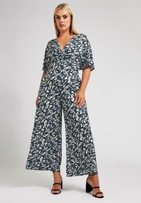 Sininen ja valkoinen kuviollinen haalari, jossa on V-mallinen kaula-aukko, lyhyet hihat, leveät lahkeet ja kireä vyötärö. Mustat korkokenkä sandaalit viimeistelevät asun.