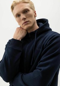 Navyblauwe ritshoodie van zachte stof. Heeft een hoge kraag, verstelbare trekkoorden en ribgebreide manchetten. Subtiele logo-detail.