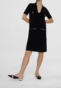 Svart kortärmad klänning med V-ringning, med två framfickor och guldkedjeaccent. I kombination med svarta och guld blockklackskor.