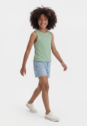 Lächendes Kind mit lockigem Haar, das einen hellgrünen ärmellosen Oberteil, Jeansshorts und weiße Turnschuhe trägt und vor weißem Hintergrund geht.