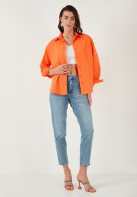 Oranje button-up shirt, witte crop top, lichtblauwe jeans en transparante hakken. Het shirt heeft lange mouwen en een ontspannen snit.