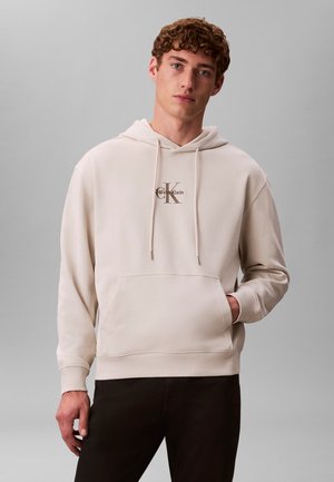 Junger Mann mit lockigen Haaren, der einen beigen Calvin Klein Hoodie und schwarze Hose trägt, vor einem einfarbigen grauen Hintergrund.