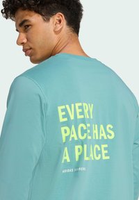 Chemise à manches longues turquoise avec du texte jaune audacieux "CHAQUE TEMPS A SA PLACE" dans le dos, fabriquée en tissu lisse avec une coupe décontractée.