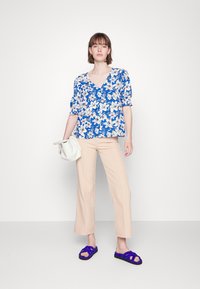 Blusa floral azul com mangas bufantes, combinada com calças largas bege e sandálias roxas. A modelo segura uma mala creme.