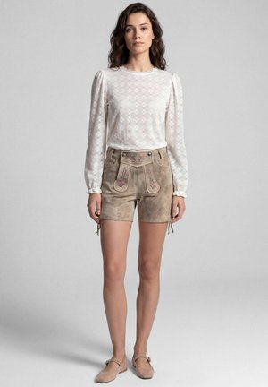 Frau steht und trägt eine weiße langärmelige Spitzenbluse, beigefarbene Wildleder-Shorts mit Blumenstickerei und beige flache Schuhe vor einem schlichten Hintergrund.