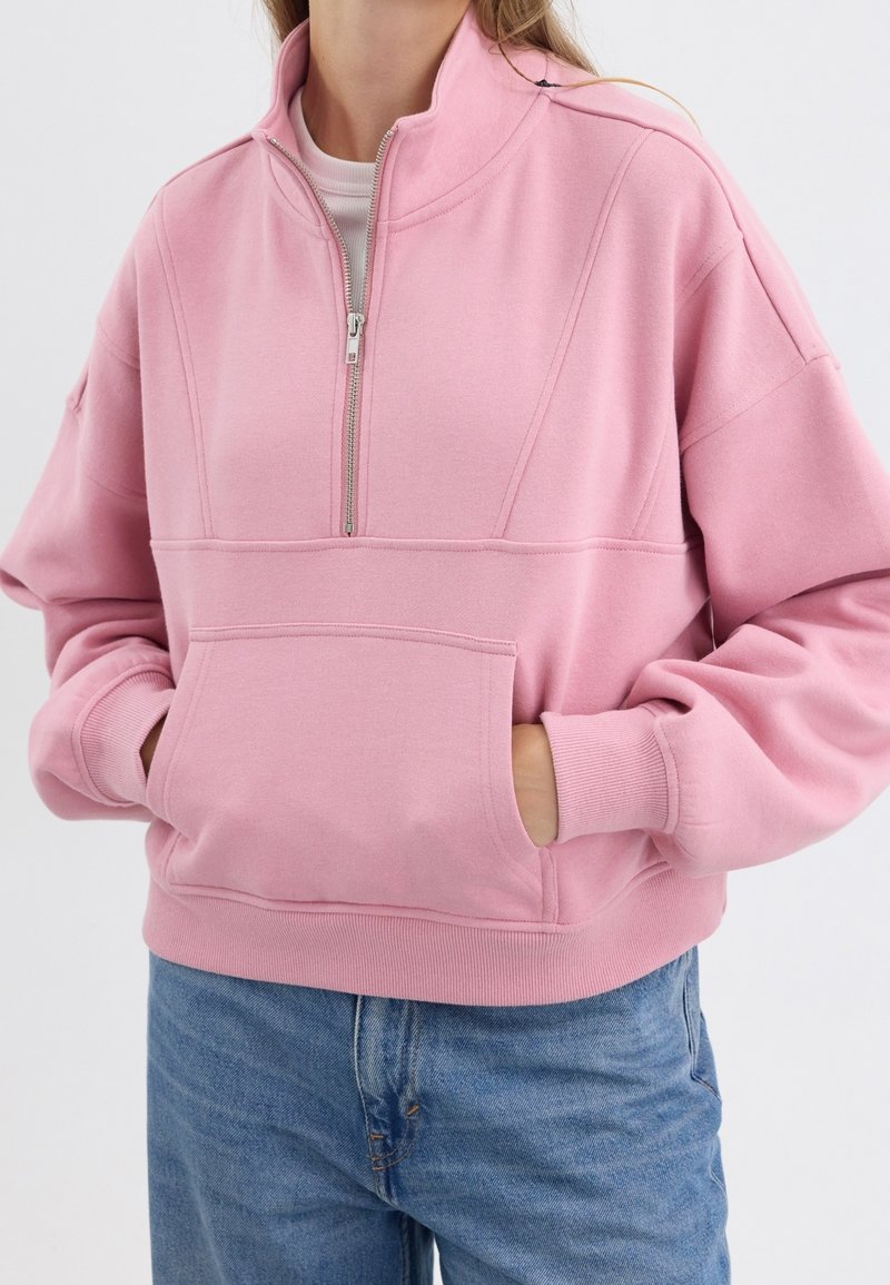 Femme portant un sweat-shirt rose clair à demi-fermeture éclair avec une poche ventrale et un jean bleu, les mains dans la poche.