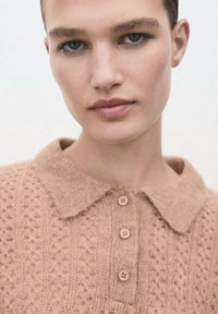 Polo de punto beige con un patrón texturizado, cuello corto y tres botones en el escote. Tejido suave con detalles intrincados.