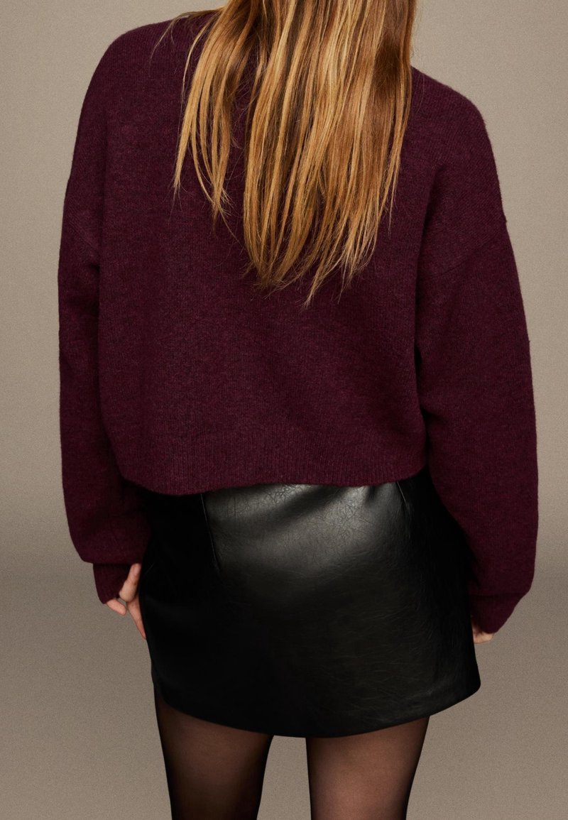Femme aux longs cheveux blonds portant un pull violet foncé et une jupe en cuir noire, debout devant un fond uni.