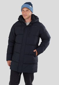 Cappotto imbottito blu Navy con design trapuntato, cerniera frontale, cappuccio regolabile e due tasche laterali. Indossato sopra un maglione grigio e pantaloni neri.