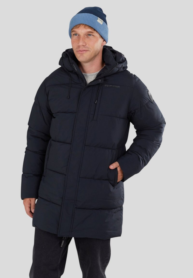 Cappotto imbottito blu Navy con design trapuntato, cerniera frontale, cappuccio regolabile e due tasche laterali. Indossato sopra un maglione grigio e pantaloni neri.