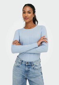 Junge Frau mit langen dunklen Haaren, die ein hellblaues geripptes Shirt und hochtaillierte blaue Jeans trägt, verschränkt selbstbewusst die Arme und lächelt.