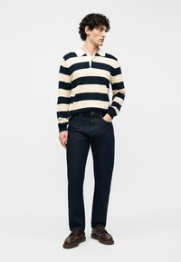 Νέος άνδρας που φοράει μπλε ναυτικό και λευκό ριγέ πουκάμισο πόλο με μακριά μανίκια, σκούρα τζιν και καφέ δερμάτινα παπούτσια, όρθιος με το ένα χέρι στην τσέπη.