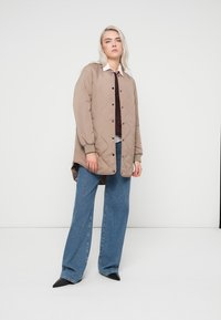 Beige izšūta jaka ar pogu priekšpusi, ribotām piedurknēm un sānu slīpēm, uzvilkta virs tumšas blūzes, kombinēta ar platām zilu džinsu biksēm un melnām kurpēm.