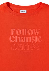 T-shirt orange vif à col rond, avec le texte en relief "Follow Change Dance" et un texte plus petit "Vis ta propre vie, poursuis ta propre voie."