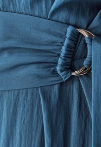 Zils audums ar gludu tekstūru, kurā ir savākta daļa un sudraba gredzena akcenti, veidojot salocītu dizainu viduklī.