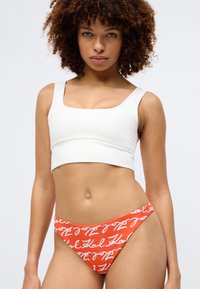 Bílý cropped top s širokými ramínky spárovaný s červenými bikinami s bílým ručně psaným vzorem. Hladký materiál, jednoduchý design.