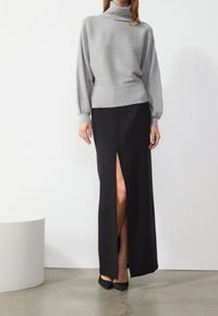 Maglione grigio a collo alto con maniche lunghe abbinato a una gonna nera maxi con uno spacco alto sulla coscia, su uno sfondo neutro.