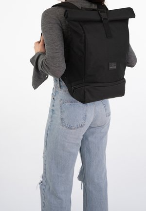 Sac à dos noir à rabat enroulable avec sangle réglable et fermeture éclair latérale, doté d'un patch logo, d'un tissu robuste et d'un design rectangulaire élégant.