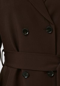 Gros plan sur un manteau croisé marron foncé avec des boutons noirs et une ceinture en tissu assorti nouée à la taille.