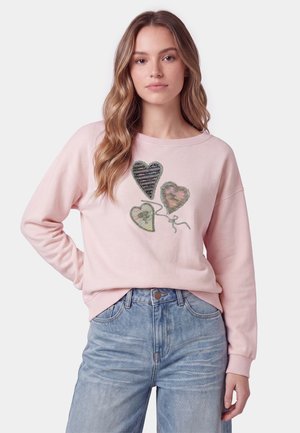 Giovane donna con capelli lunghi e mossi indossa una felpa rosa chiaro con tre disegni a forma di palloncino a cuore e jeans blu a vita alta.