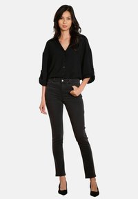 Mujer con cabello largo y oscuro, vestida con una camisa negra de botón suelta, jeans skinny negros de cintura alta y tacones de punta negros, de pie con confianza.