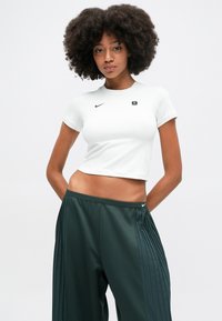 T-shirt branca cropped com mangas curtas, apresentando um pequeno logotipo preto da Nike. Combinada com calças pregueadas verde-escuras com um cós elástico.