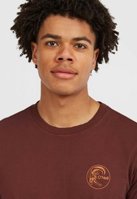 Bordeaux kleurige langemouwen T-shirt met geribde halslijn en logo met een oranje grafisch ontwerp op het linker borstgebied. Zacht katoen materiaal.
