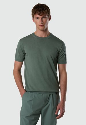 Mann mit kurzen lockigen Haaren, der ein einfaches grünes kurzärmeliges Hemd und passende grüne Hosen trägt, mit den Händen in den Taschen stehend.