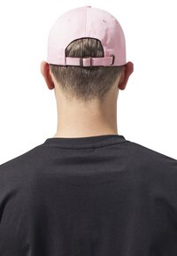 Roze katoenen pet met een verstelbare band aan de achterkant, gedragen door een persoon die van zich af kijkt, gecombineerd met een zwart T-shirt.