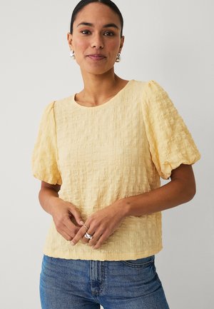 Kvinde iført en struktureret, lysegul bluse med puffærmer og blå jeans, med sølvøreringe og ringe, står mod en ensfarvet baggrund.