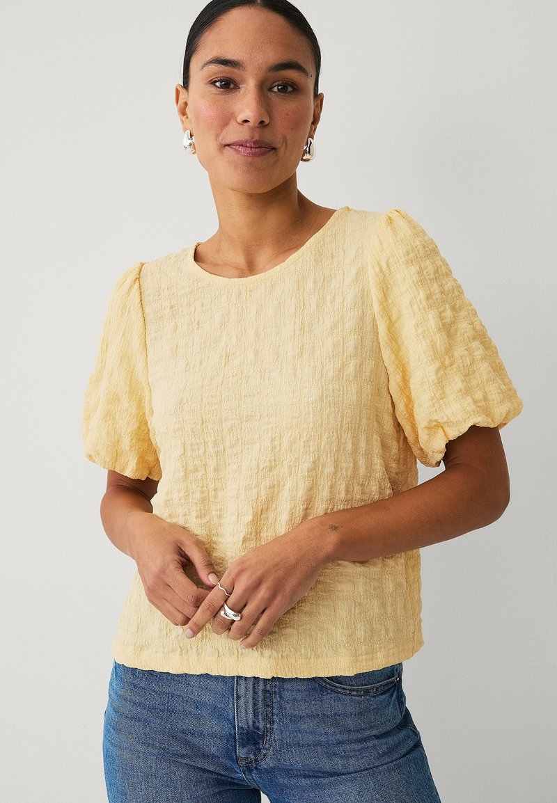 Mujer con blusa amarilla clara de manga abullonada y textura visible, y jeans azules, luciendo pendientes y anillos plateados, de pie contra un fondo liso.