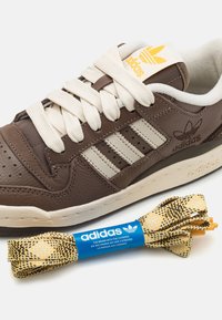 Brown leather sneaker with cream accents, three white stripes, and beige laces. Includes yellow and black patterned shoelaces with blue label.