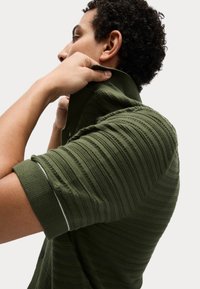 Polo de manga corta verde con rayas texturizadas, collar y puños acanalados con detalles en blanco. La tela parece suave y ligera.
