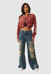 Vrouw in een cropped rode geruite jas, zwarte graphic crop top, versleten wijd gesneden jeans en witte sneakers, staand tegen een effen achtergrond.