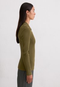 Olijfgroen geribbeld polo met lange mouwen en kraag, getailleerd model en textielstructuur. Gecombineerd met grijze broek, weergegeven in profielweergave.