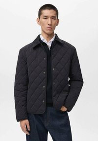 Chaqueta acolchada negra con cuello, que presenta patrones de diamantes, botones a presión y un diseño ajustado, llevada sobre un suéter oscuro y una camisa.