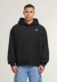 Sudadera de algodón negra con un bolsillo frontal, que presenta un pequeño logo blanco en el pecho y una capucha con cordón. El modelo lleva pantalones cargo en azul claro.