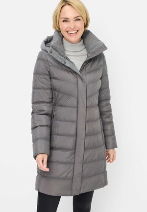 Lächelnde Frau trägt einen langen grauen Steppmantel mit hohem Kragen und seitlichen Taschen über einem hellgrauen Rollkragenpullover.