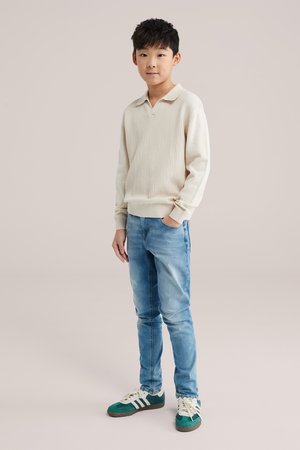 Poiss seisab neutraalse taustaga, üks käsi taskus, kandes beežist kampsunit, hele siniseid teksaseid ja rohevalgeid tenniseid.