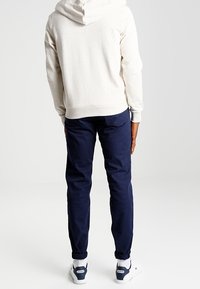Sudadera con capucha beige de tejido texturizado, combinada con pantalones azul marino. Zapatillas blancas con detalles azules completan el atuendo visto desde la parte trasera.