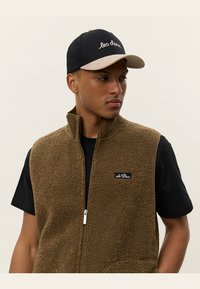 Brown fleece vest with a zip front and side pockets, worn over a black t-shirt. A black cap with a tan brim and embroidered text is also visible.