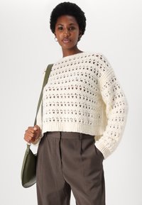 Krém színű, kötött pulóver nyitott mintával, cropped fazonnal és bordás hemöléssel. A modell zöld táskát tart és barna nadrágot visel.