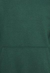 Felpa verde in cotone con una texture liscia, tasca a marsupio frontale e dettagli cuciti lungo le cuciture. Design semplice e pullover.