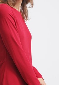 Punainen pitkähihaista mekkoa, jossa on muotoiltu vartalo ja levenevä helma. Kangas näyttää sileältä ja siinä on vähäistä kiiltoa; minimalistinen muotoilu ilman näkyviä kuvioita.