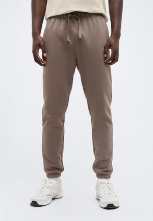 Homme portant un pantalon de jogging beige à cordon de serrage et des baskets blanches, debout devant un fond clair uni.