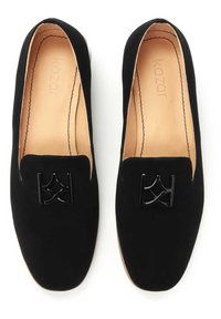 Melni zamšaini loafers ar gludu tekstūru, ar spīdīgu melnu metāla emblemu priekšpusē un bežīgas ādas iekšpusi.