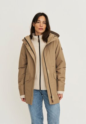 Vrouw draagt een beige jas met capuchon over een crème sweater met rits en een blauwe spijkerbroek, staand tegen een eenvoudige lichte achtergrond.