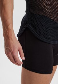 Sort mesh tanktop med en løs pasform og rundet kant, parret med tætsiddende sorte shorts. Synlig hånd på tanktoppen.