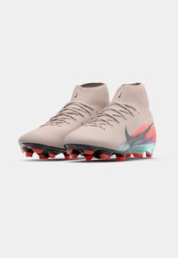 Ζευγάρι μπεζ ψηλές ποδοσφαιρικές μπότες με μαύρες και κόκκινες τακουνιές από καουτσούκ και μια μεγάλη σκούρα γκρι νικέλ σλόγκαν στις πλευρές.