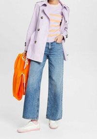 Lavenderfarbener Trenchcoat, gestreifter Pullover in Lavendel und Pfirsich, weit geschnittene Jeans, weiße Sneakers mit pinken Akzenten und hält einen orangenen Kapuzenpulli.